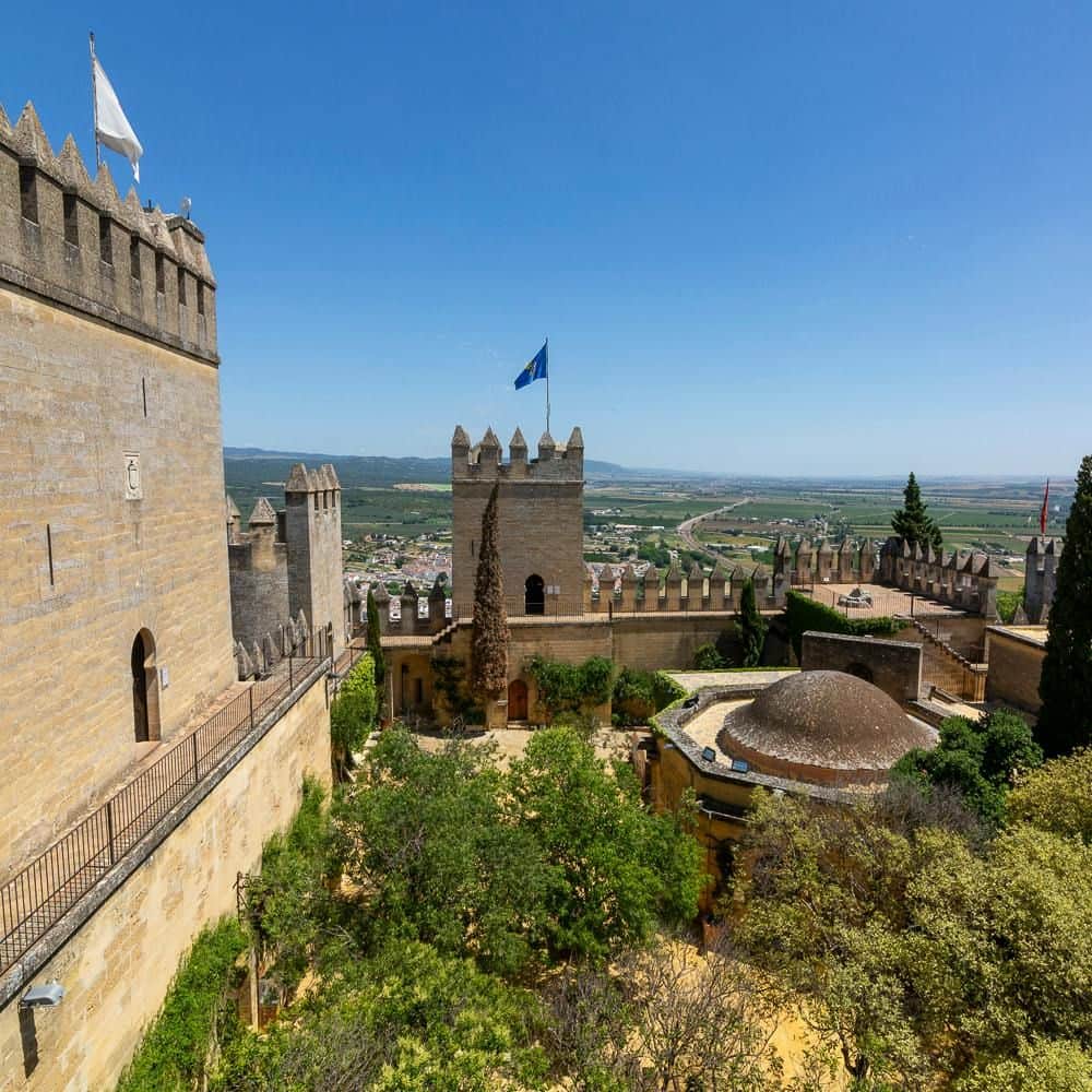 Almodóvar Castle: Entry Ticket Stone fortress with crenellated walls and a flag on a central tower, surrounded by greenery and overlooking a vast rural landscape.