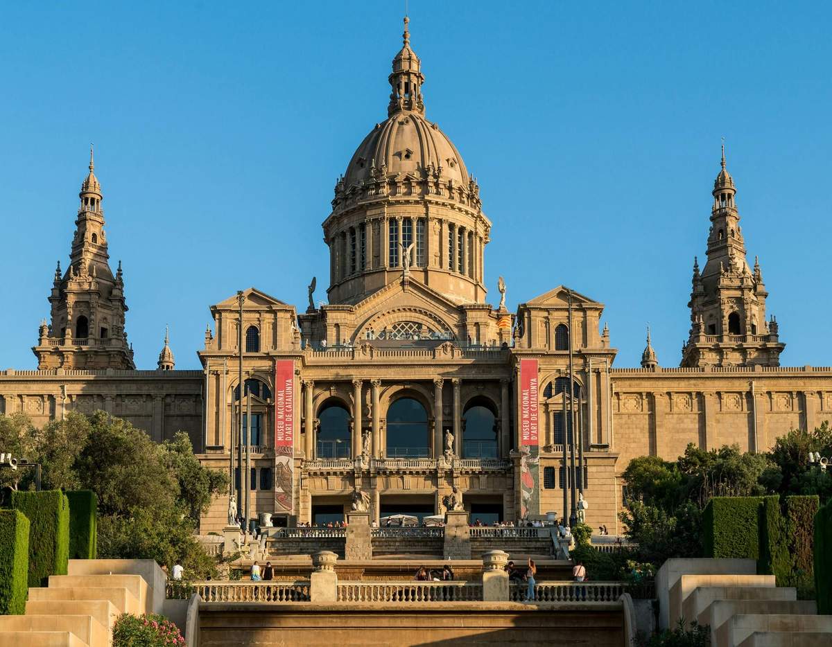 Museu Nacional d’Art de Catalunya: Skip The Line Ticket Ornate building with large dome and towers, featuring arched windows and flanked by two red banners, under a clear blue sky.