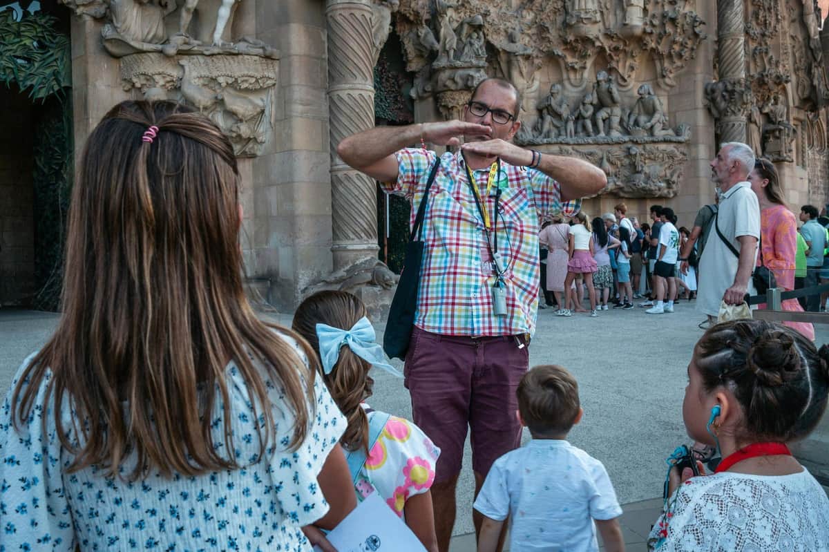 A tour guide in a plaid shirt gestures while speaking to a group of children outside an intricately detailed stone building.