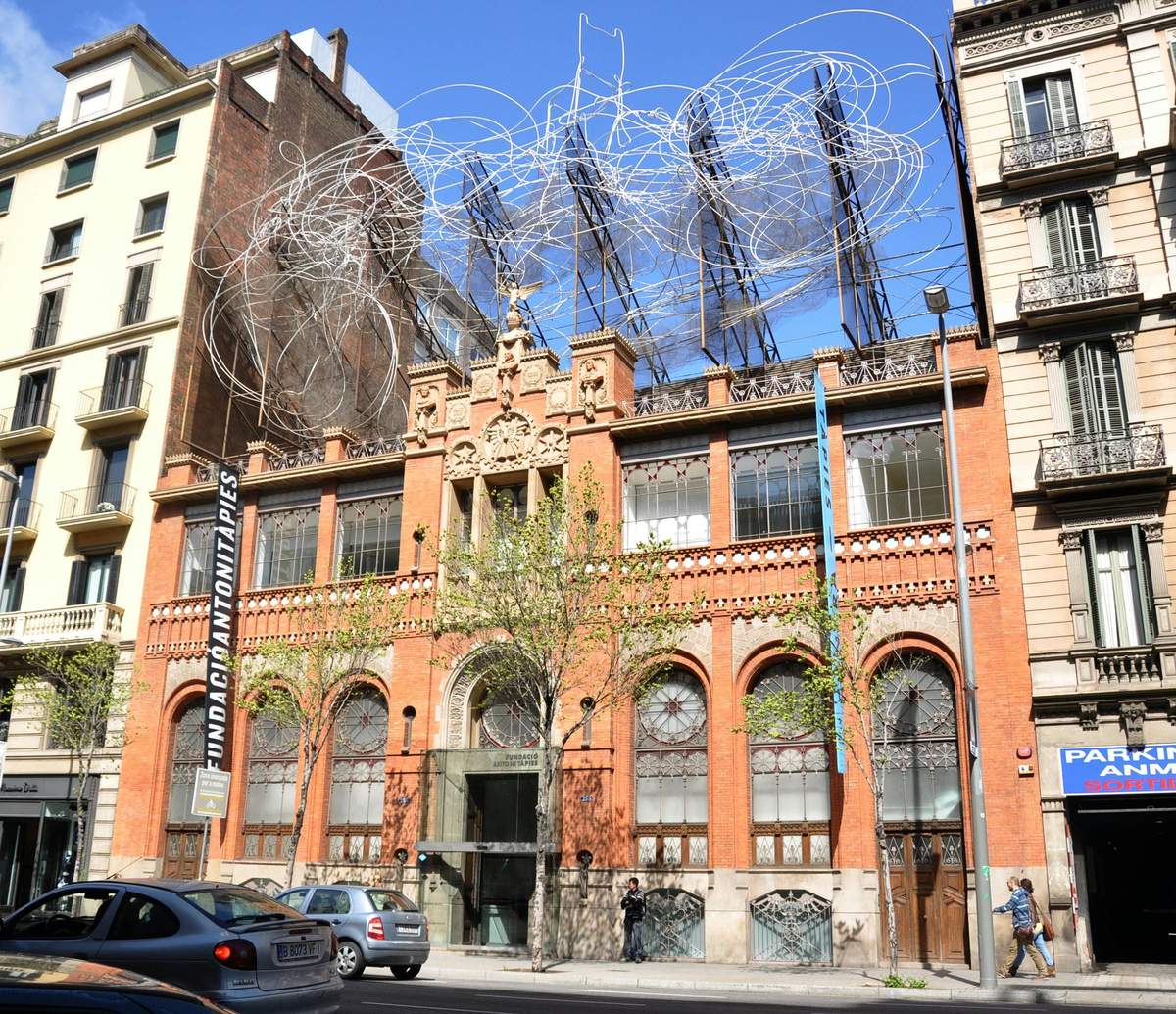 Tàpies Museum: Skip The Line Ticket A red-brick building with large arched windows, artistic wire sculptures on the roof, and cars parked in front on a sunny day.