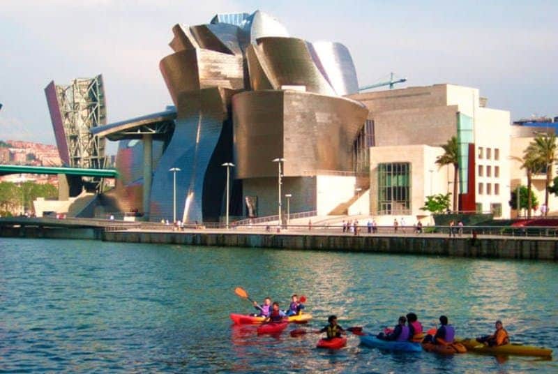 Guggenheim Museum Bilbao: Skip The Line Ticket + Guided Tour People kayaking in a river near a modern, metallic building with a unique architectural design under a clear sky.