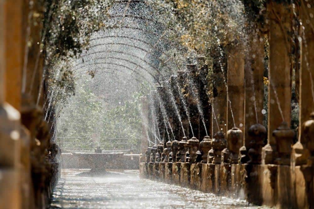 Jardines de Alfabia: Skip The Line Ticket Arched walkway with water spraying from fountains on both sides, creating a misty effect. Lush green foliage in the background.
