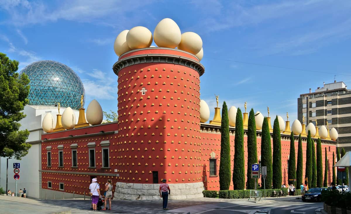 Dalí Theatre and Museum: Fast Track Ticket Red brick building with large eggs on top and dotted decorative elements, surrounded by tall trees. People stand at the corner.