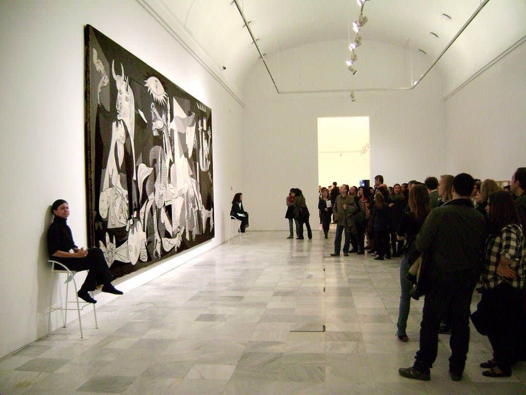 Museo Reina Sofía: Skip The Line Ticket Visitors observe Picasso's "Guernica" painting in a museum. Guides sit on chairs near the artwork.