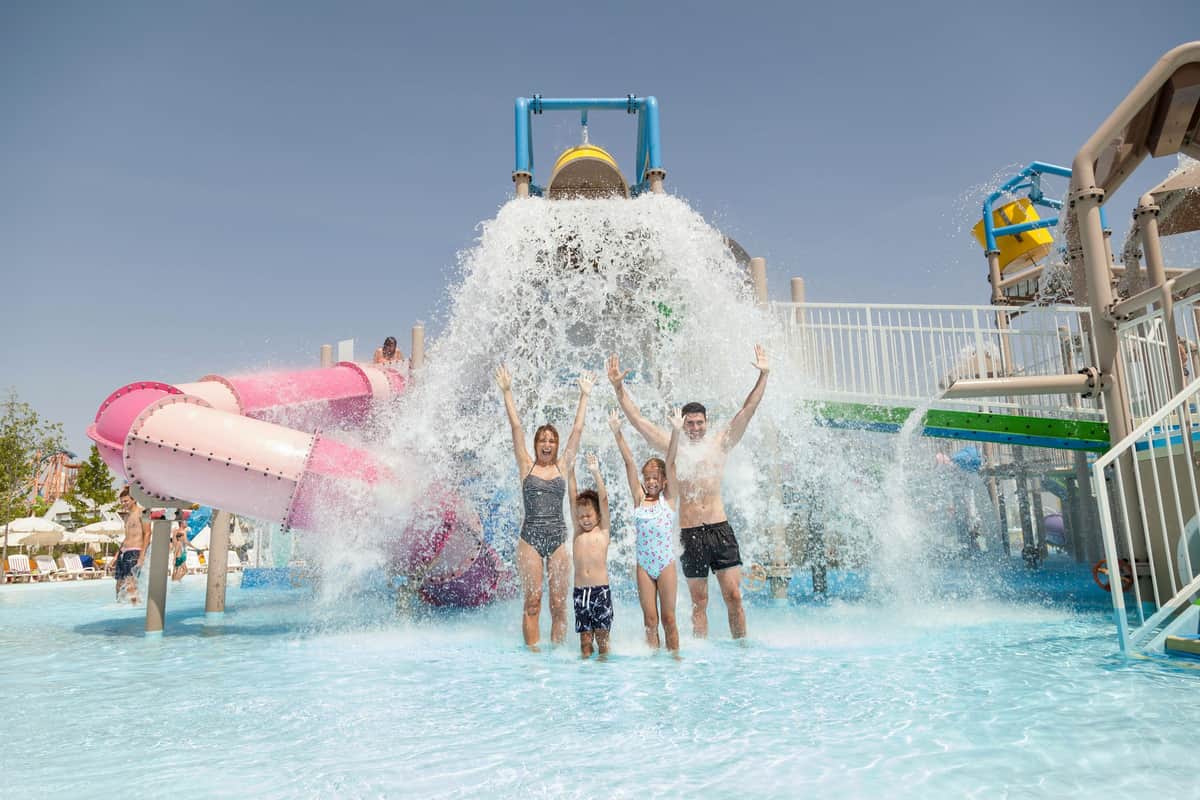 A family of four stands under a large water bucket splash at a water park, smiling and raising their arms as water cascades down.