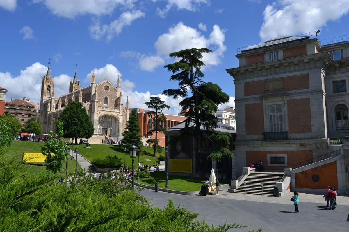 A historic church, a tree, and a museum building with stairs in a square under a partly cloudy blue sky.