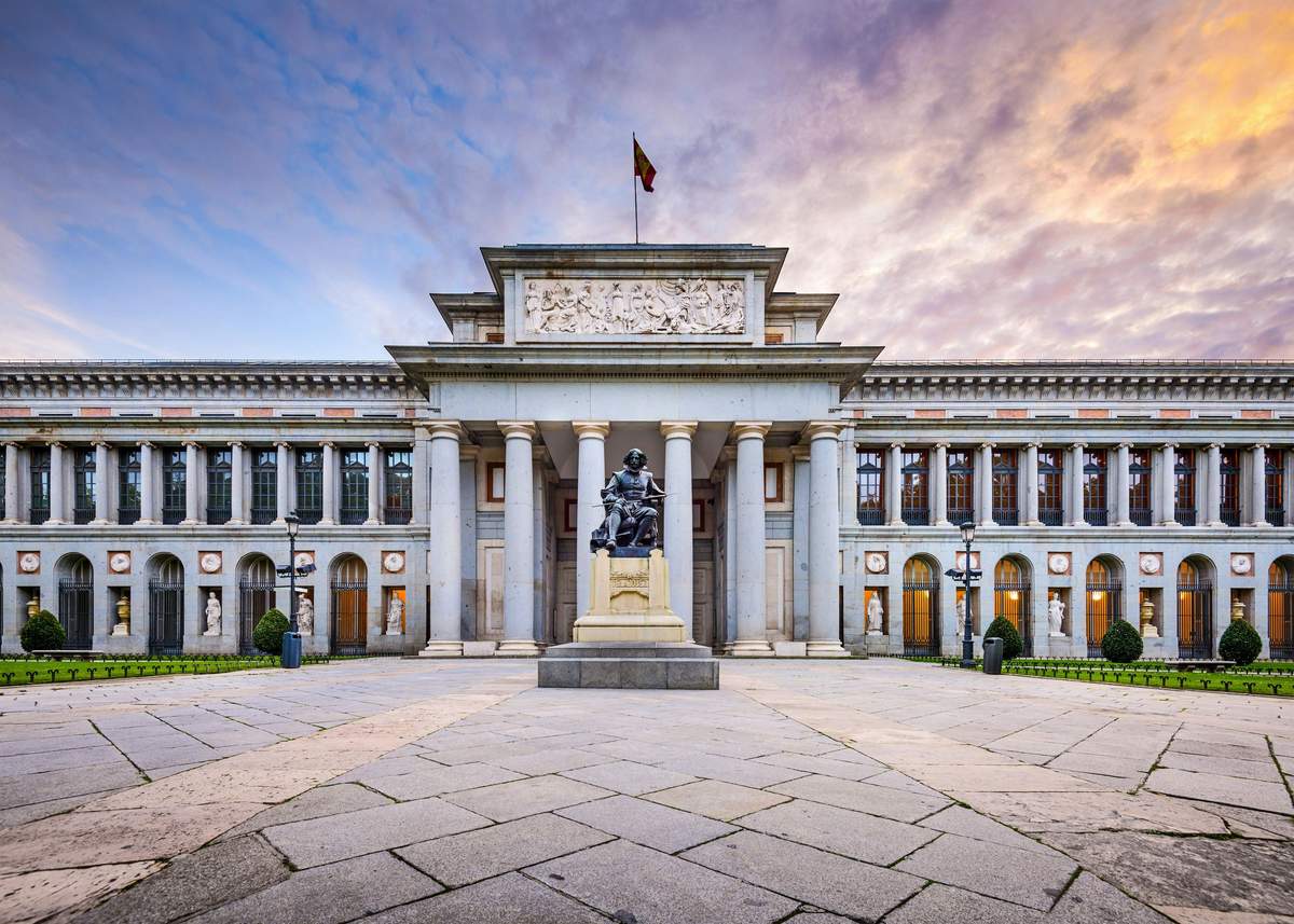 Prado Museum: Skip The Line Ticket + Guided Tour A neoclassical building with columns, a statue in front, and a flag on top, under a colourful sunset sky.
