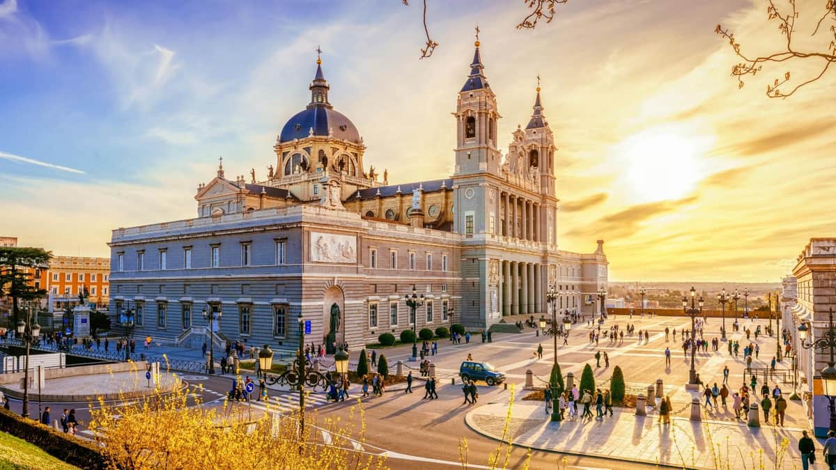 Royal Palace Madrid and Cathedral of La Almudena: Skip-The-Line & Guided Tour A grand building with domes and towers under a sunset sky, surrounded by people, cars, and manicured gardens.