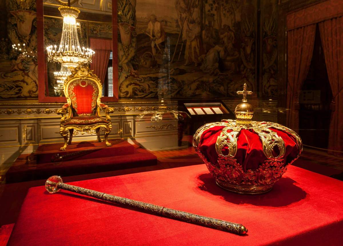 Royal Palace of Madrid: Fast Track Entry An ornate crown and scepter on a red velvet surface in a room with a gilded throne and a chandelier.