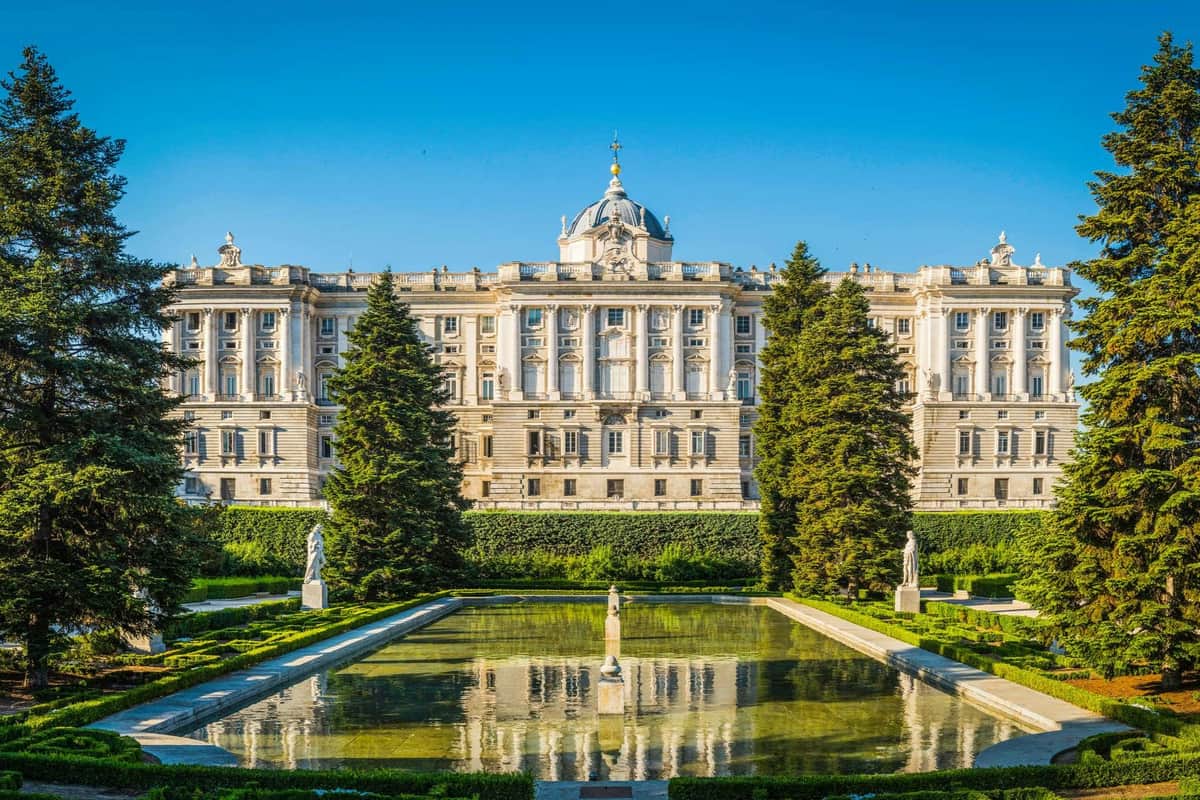 Royal Palace of Madrid: Skip The Line Ticket + Small Group Guided Tour A grand palace with a domed roof, seen behind manicured gardens, statues, and a reflecting pool under a clear blue sky.