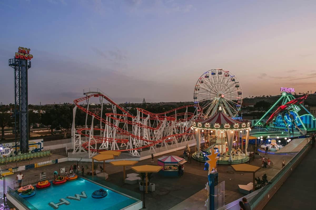 Holidayworld Wooland Fun Park: Fast Track An amusement park at dusk with a rollercoaster, Ferris wheel, carousel, bumper boats, and colorful lights illuminating the area.