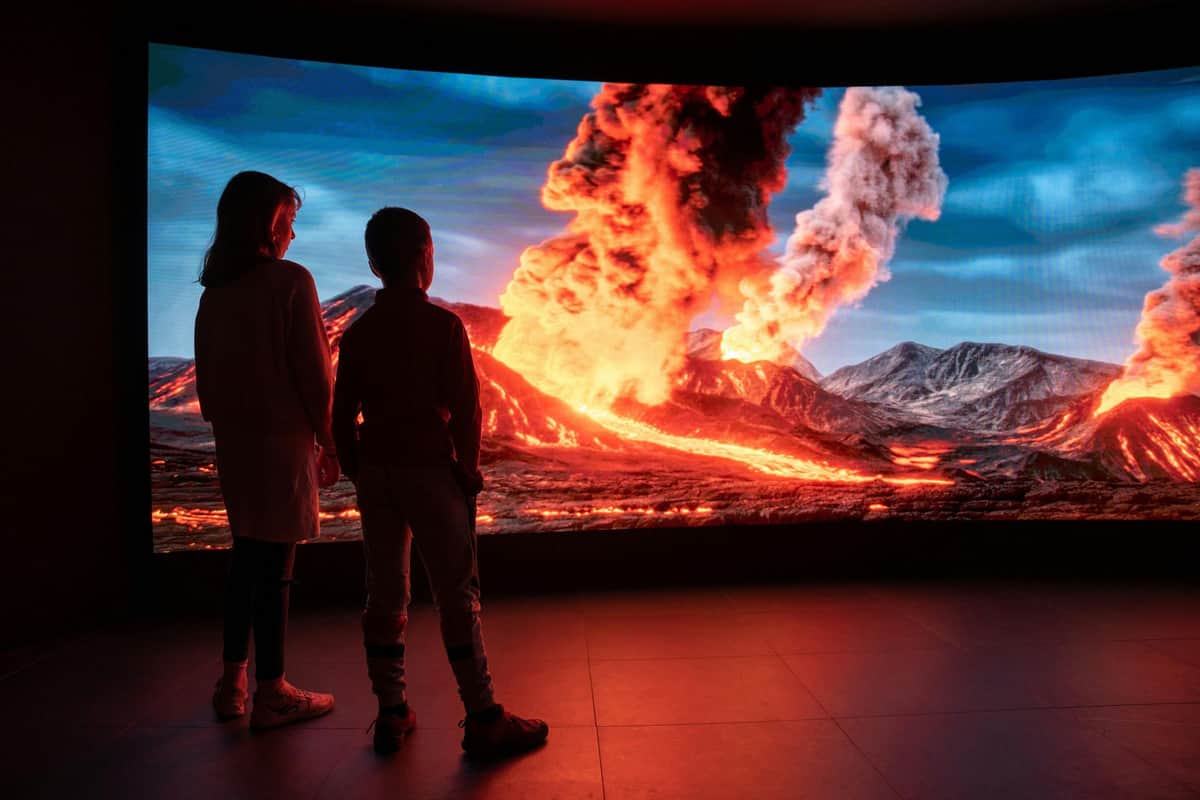 Espai Cràter: Skip The Line Ticket Two children stand in front of a large screen showing a dramatic volcanic eruption with flowing lava and smoke under a blue sky.