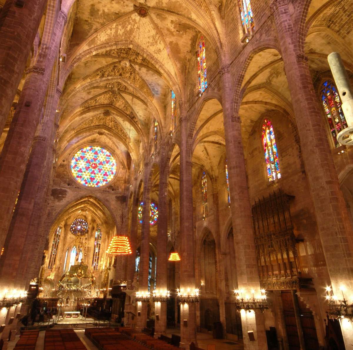 Mallorca Cathedral Interior of a grand cathedral with high arched ceilings, intricate stained glass windows, and hanging chandeliers.