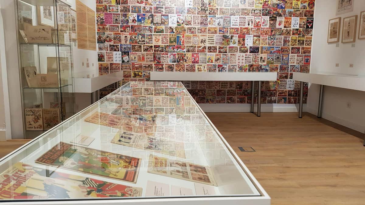 Museu del Còmic i la Il·lustració: Skip The Line Ticket A display case with comic book pages, surrounded by walls covered in comic book artwork, in a well-lit exhibition space.