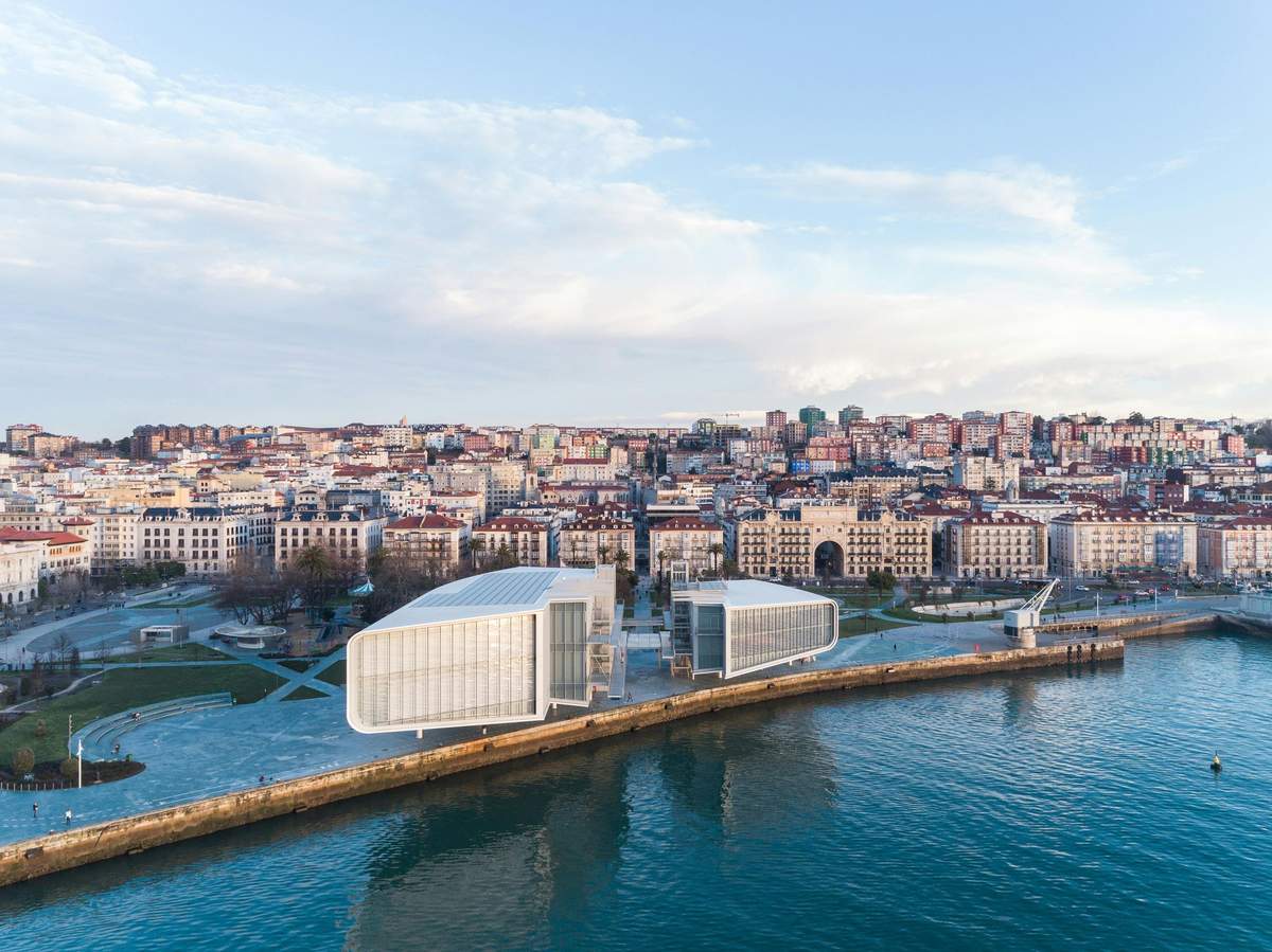 Modern waterfront buildings near a cityscape with densely packed buildings, a park area, and a clear blue sky.