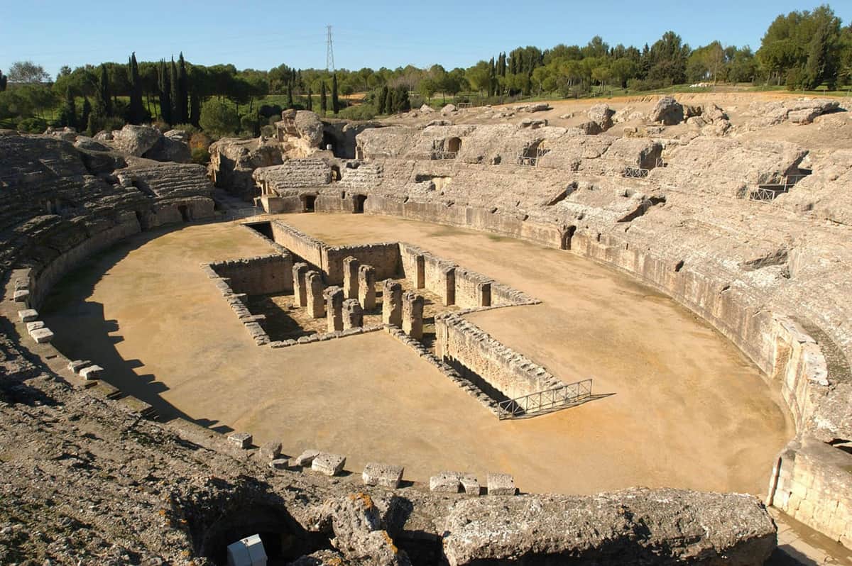 Game of Thrones at Itálica: Guided Visit & Skip The Line Aerial view of an ancient amphitheater with stone ruins in the center, surrounded by tree-covered hills.