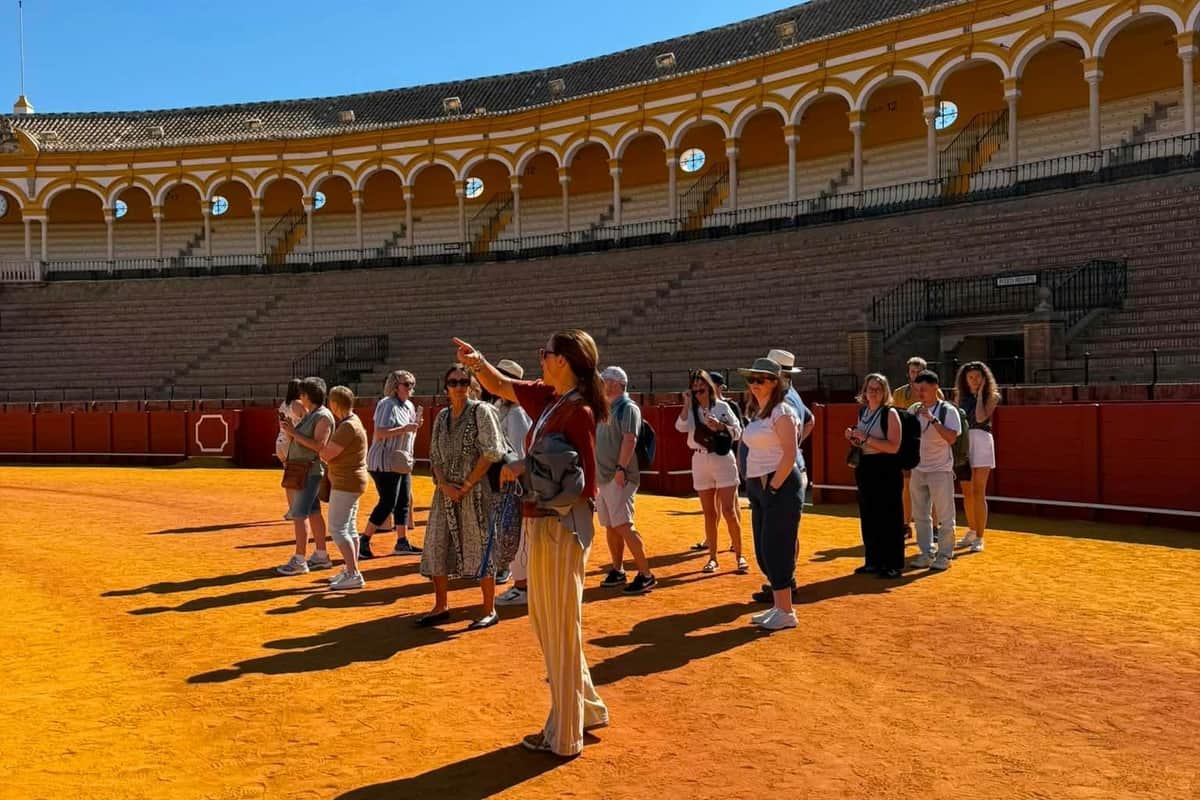 Real Maestranza: Guided Tour A group of people standing on the floor of an arena with tiered seating in the background, beneath a clear blue sky.