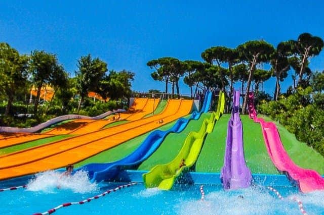 Illa Fantasia Waterpark: Skip The Line Ticket Colourful water slides with people sliding into a pool, surrounded by trees and greenery under a clear blue sky.