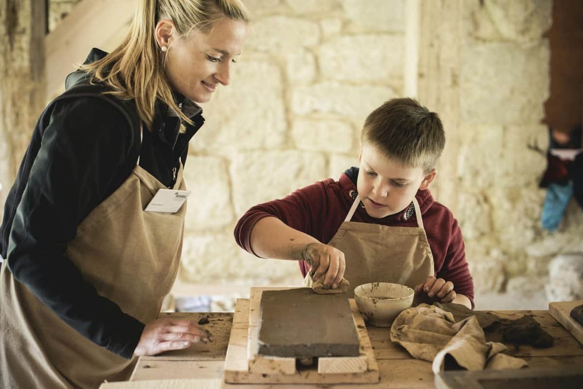 Open-Air Museum Ballenberg: Skip The Line Ticket A woman and a boy, both wearing aprons, work with clay on a wooden table in a rustic setting.