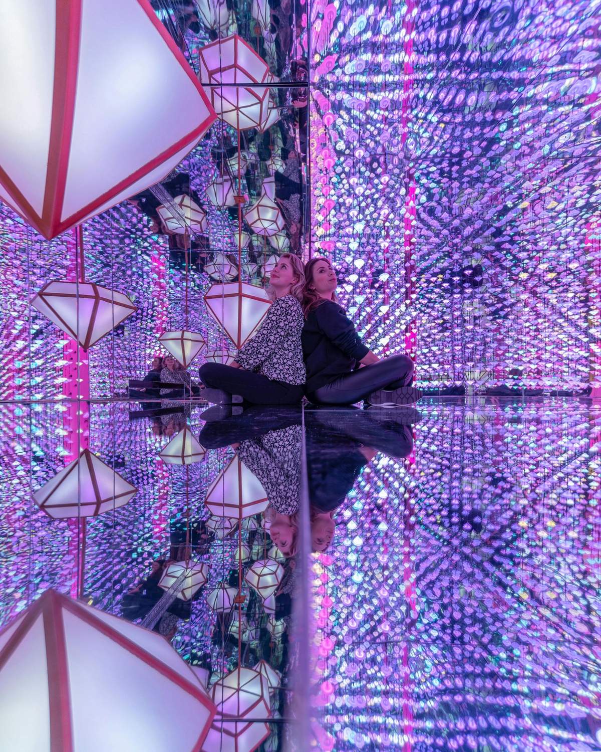 The Upside Down + Moco Museum Two women sit back-to-back on a reflective floor surrounded by mirrors and colorful LED lights, with hanging geometric lamps above.
