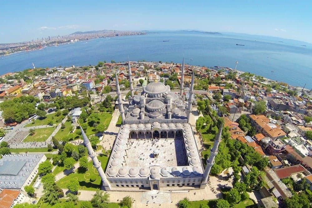 The Blue Mosque & Hagia Sophia: Small Group Guided Tour Aerial view of a large mosque with multiple domes and minarets, surrounded by trees and buildings, near a body of water.