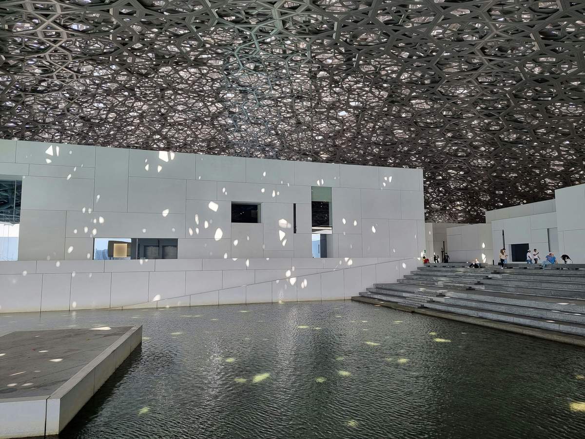 Louvre Abu Dhabi: Skip The Line Ticket Modern building with geometric-patterned ceiling, white walls, water feature, and steps. Few people sit on the steps.