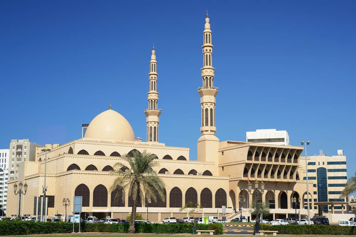 Sharjah Heritage Museum: Skip The Line & Sharjah City Tour from Dubai Large mosque with two tall minarets, a dome, arched windows, and a palm tree in the foreground under a clear blue sky.