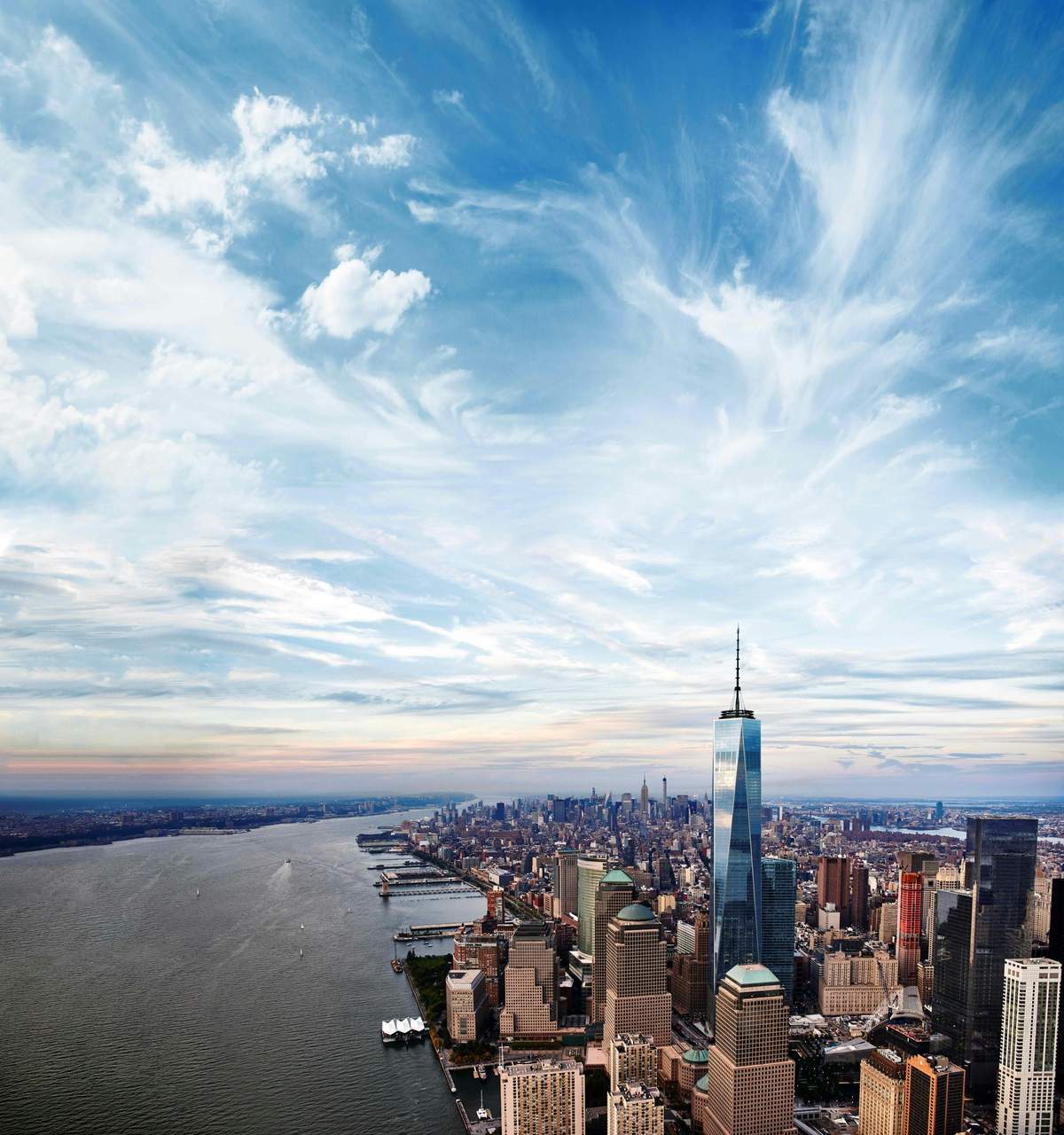 Aerial view of downtown Manhattan with skyscrapers, including One World Trade Center, and the Hudson River under a partly cloudy sky.