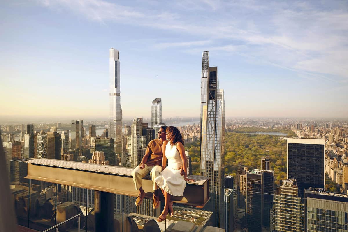 A couple sits on a beam atop a skyscraper, overlooking a cityscape with tall buildings and a distant park under a blue sky.