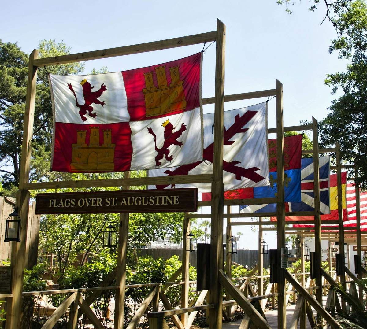 Colonial Experience: Skip The Line Colorful historic flags hang outdoors on wooden frames with a sign below reading "Flags Over St. Augustine." Trees and foliage surround the area.