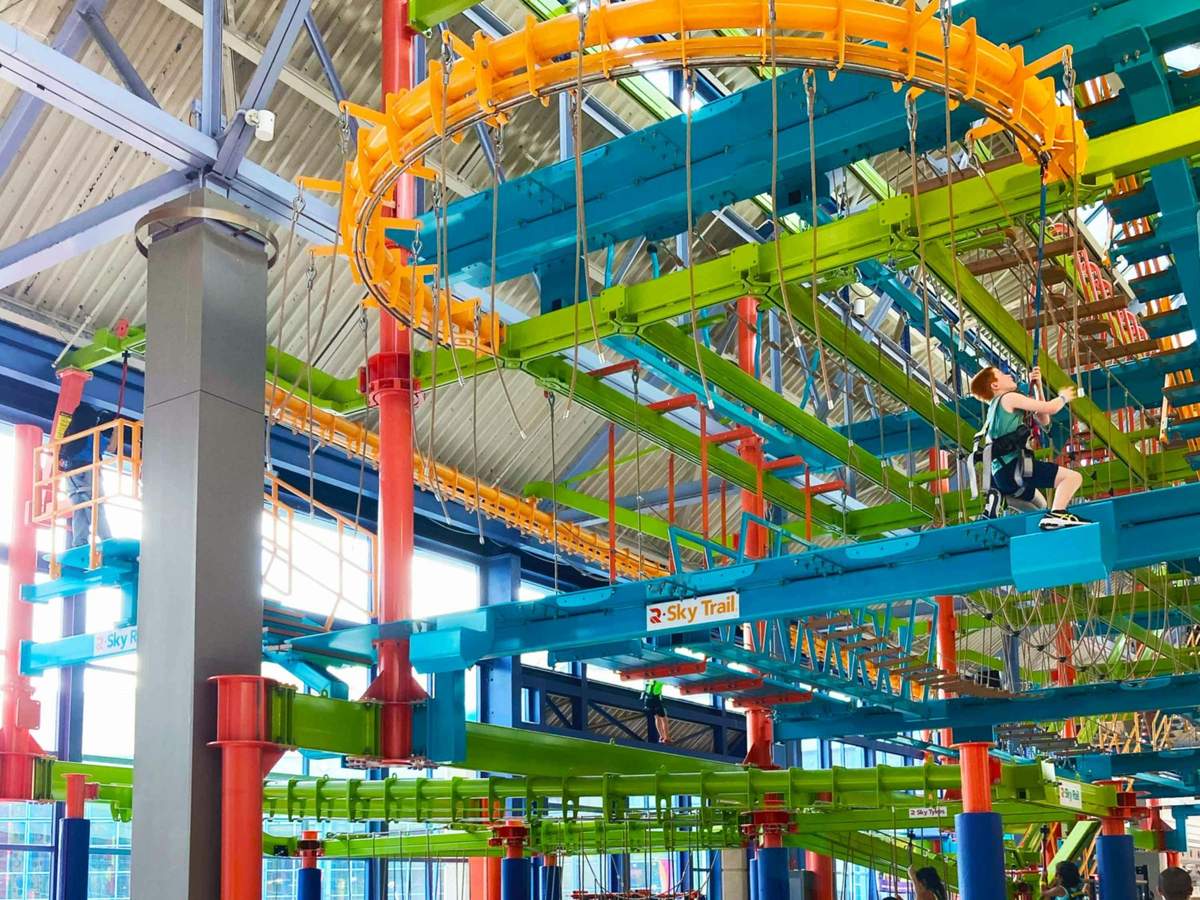 LiggettVille San Antonio: Fast Track + High Ropes Course, Zip Rail & Wall Climb Indoor ropes course with colorful obstacles, safety harnesses, and ropes. A "Sky Trail" sign is visible, and one person is on the course.