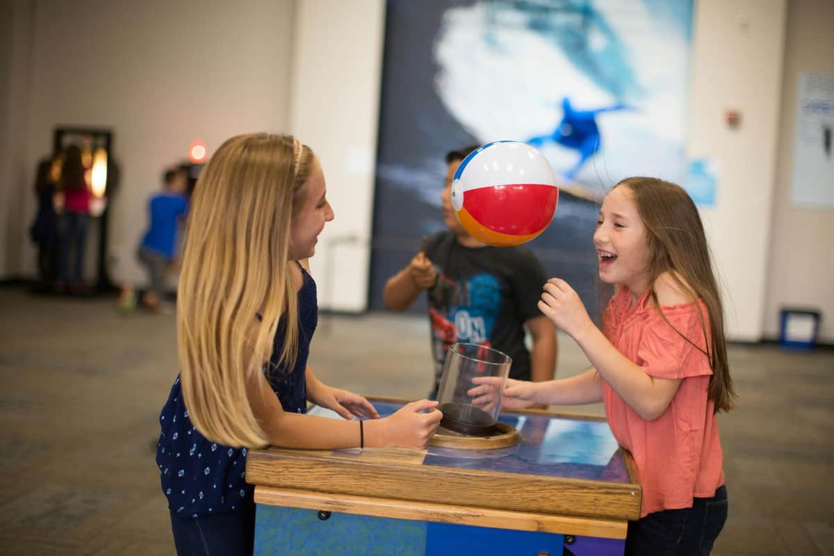 The Fleet Science Center: Fast Track Ticket Two girls are playing with a floating beach ball in a science exhibit, with a blurred surfer image in the background.