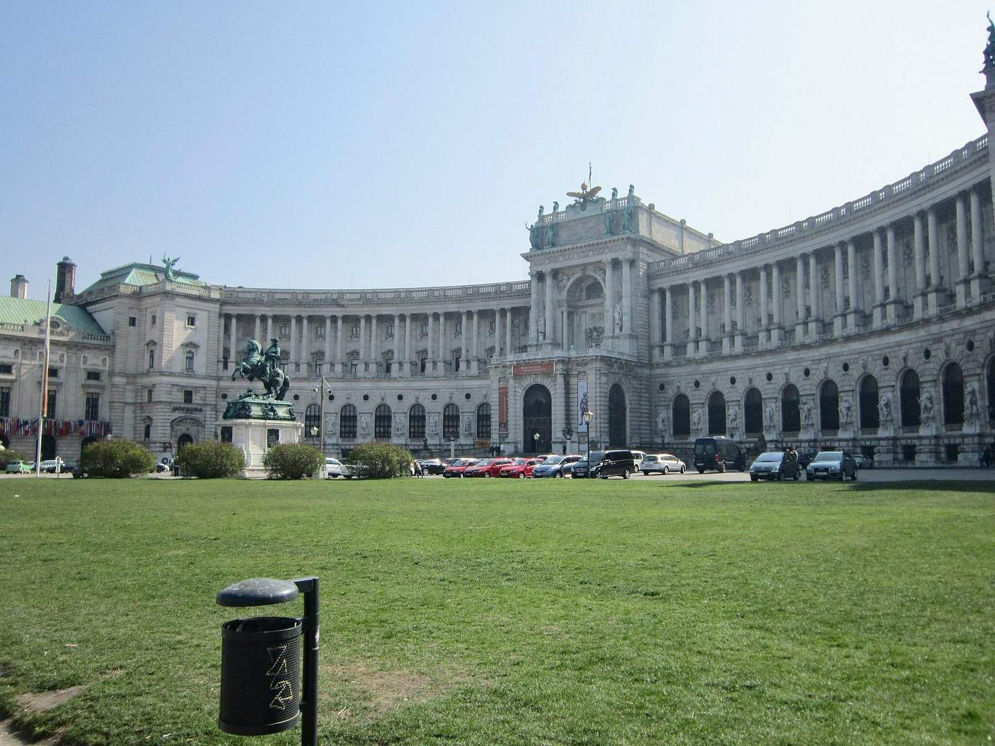 Hofburg Palace Grand building with curved facade, statue of rider on horse in front, cars parked along the street, green lawn and trash can.