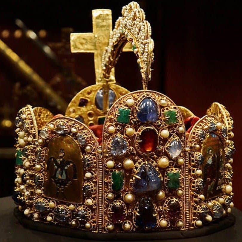 Imperial Treasury An ornate golden crown adorned with jewels, pearls, religious icons, and a cross on top, displayed against a dark background.
