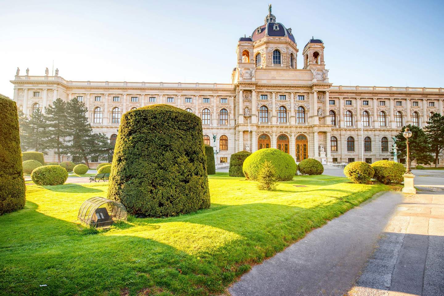 Kunsthistorisches Museum A large, historic building with arched windows, a dome, and neatly trimmed hedges in a sunlit garden area.