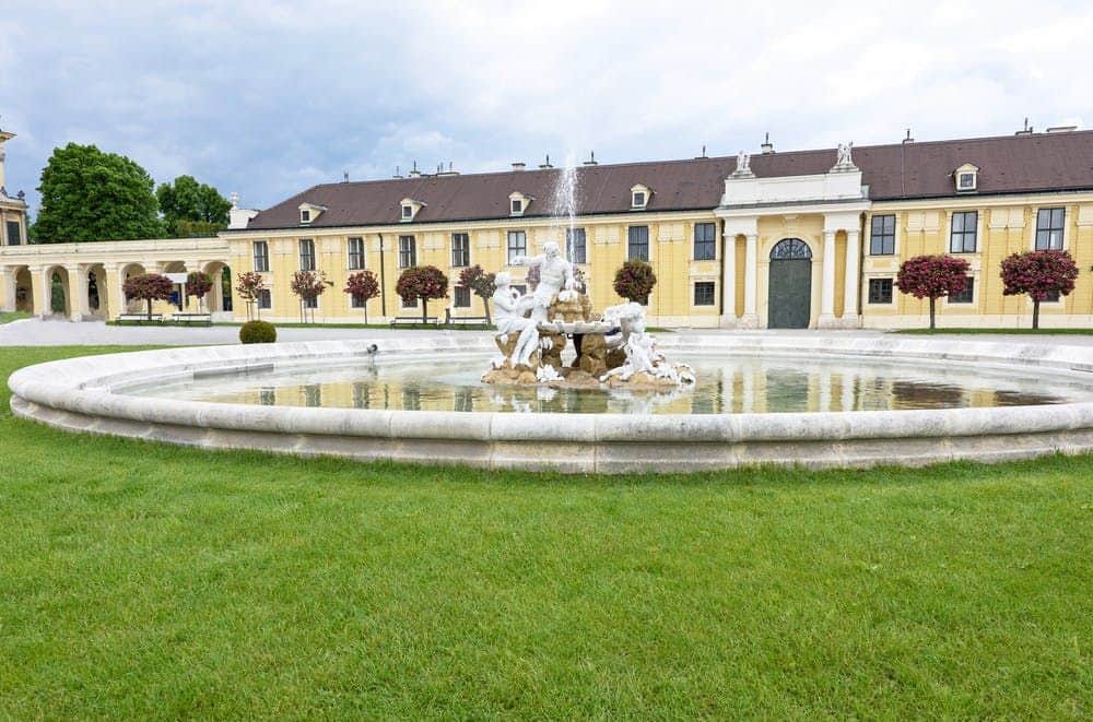 Schönbrunn Palace: Concert at the Orangerie Ornate fountain with statues in a landscaped garden, with a long, yellow building and neatly trimmed trees in the background.