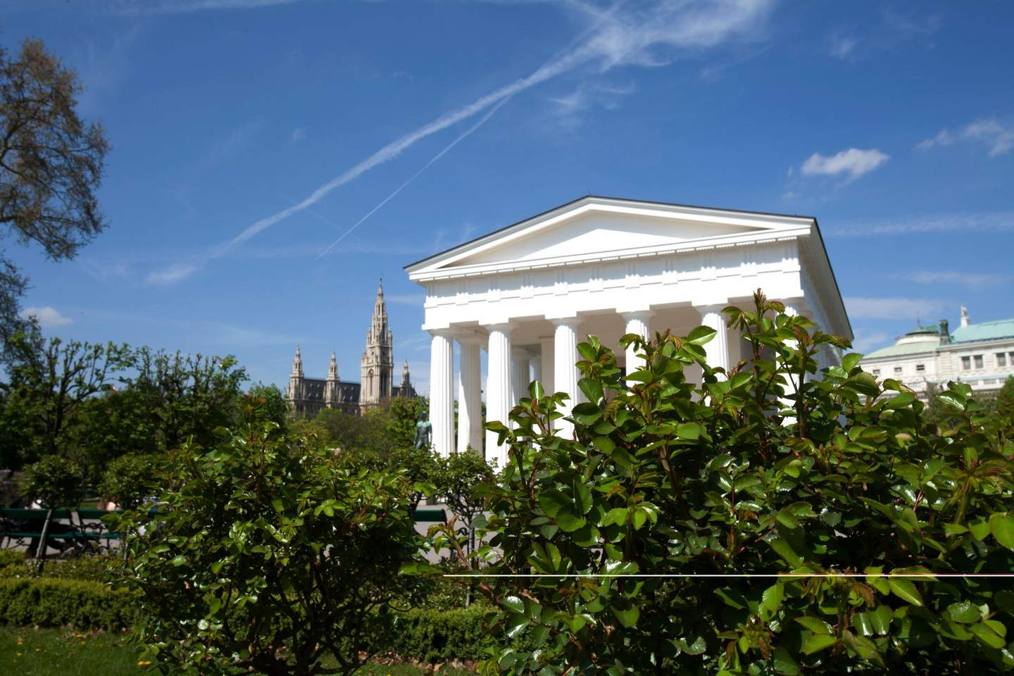 White neoclassical building with columns surrounded by greenery, distant cathedral spires, under a blue sky with wispy clouds.
