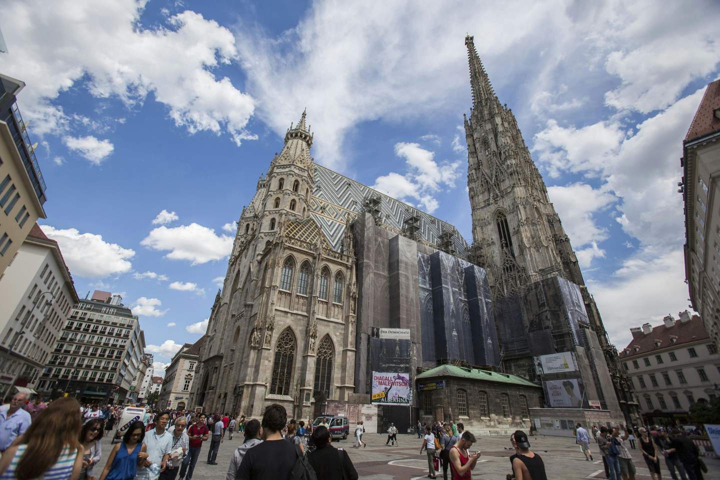 Cattedrale di Santo Stefano Folla di persone che cammina vicino a una cattedrale in stile gotico con un'alta guglia, coperta in parte per lavori di ristrutturazione, sotto un cielo azzurro con nuvole.