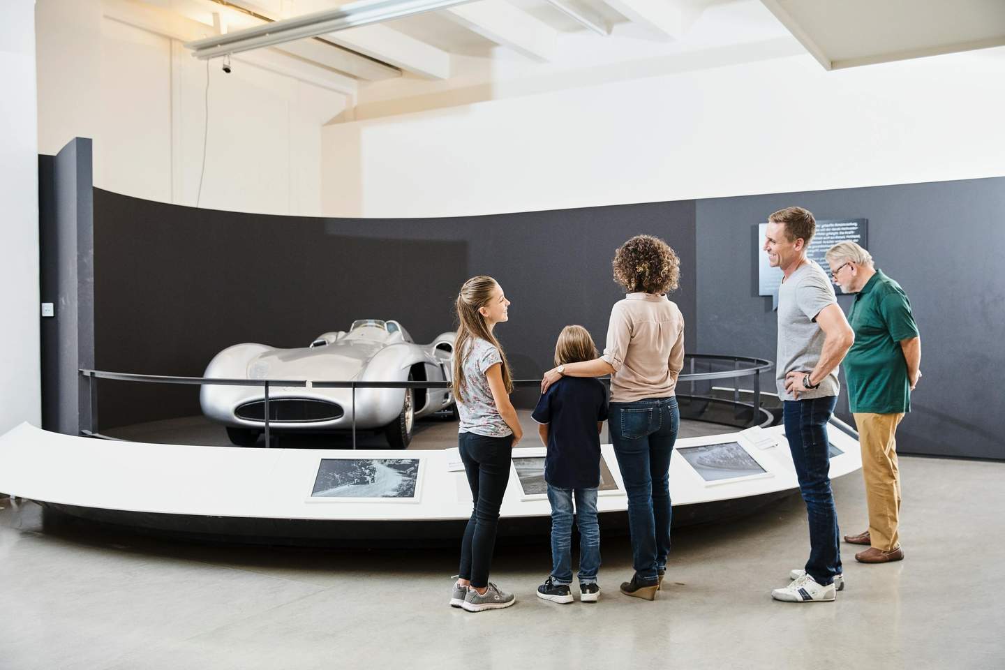 Vienna Technical Museum Five people admiring a silver vintage race car displayed in a museum with informational plaques in front.