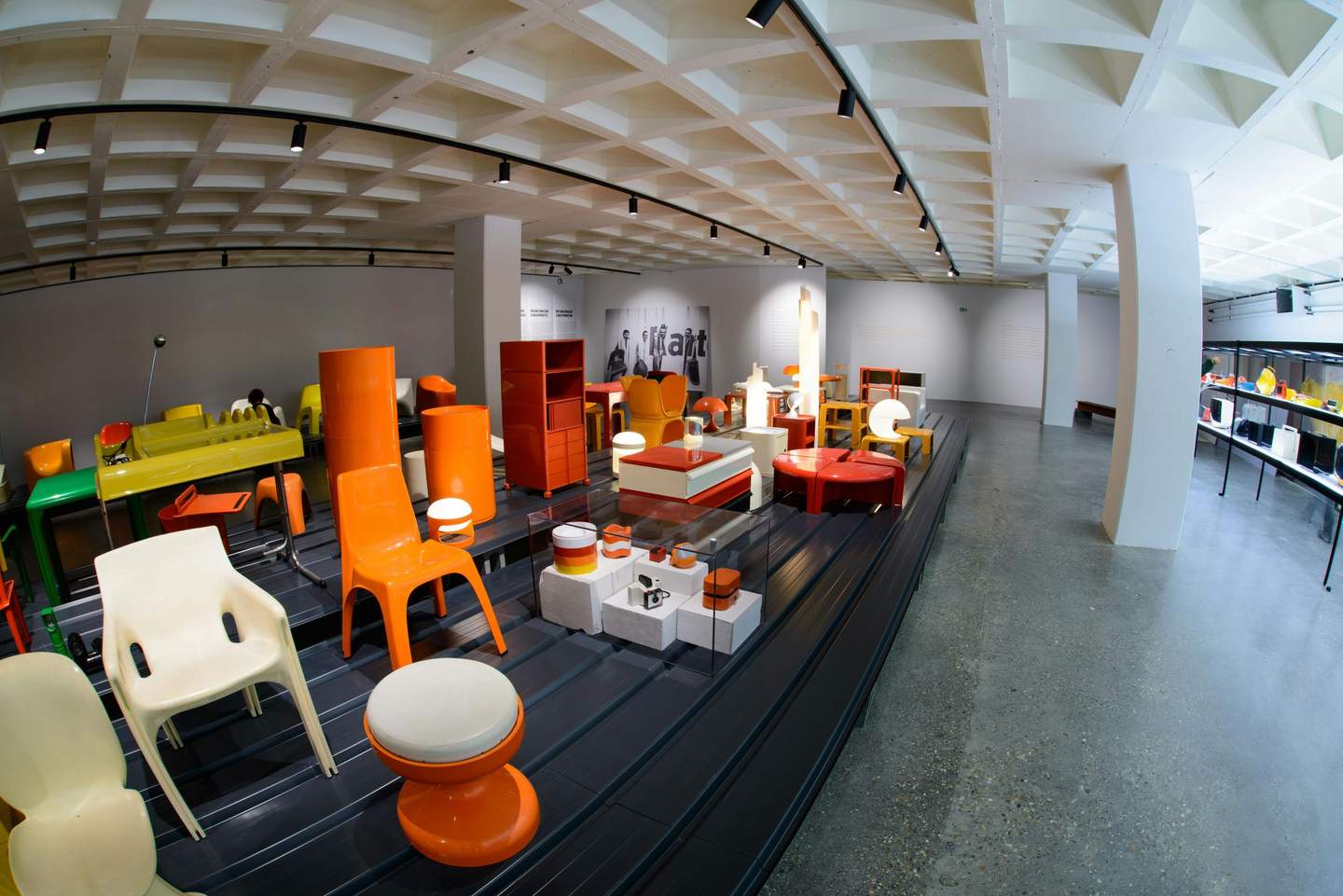 Exhibit room showcasing various colorful plastic furniture pieces and lamps, under a coffered ceiling with track lighting.