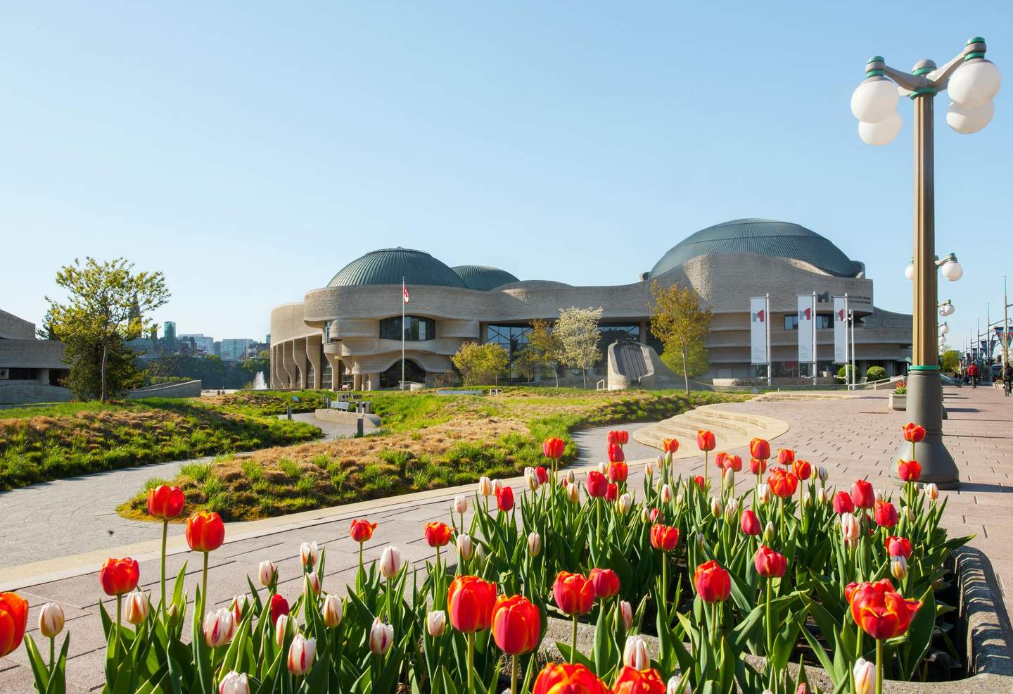 A modern building with curved architecture, a Canadian flag, red and white tulips, and a pathway with street lamps on a sunny day.