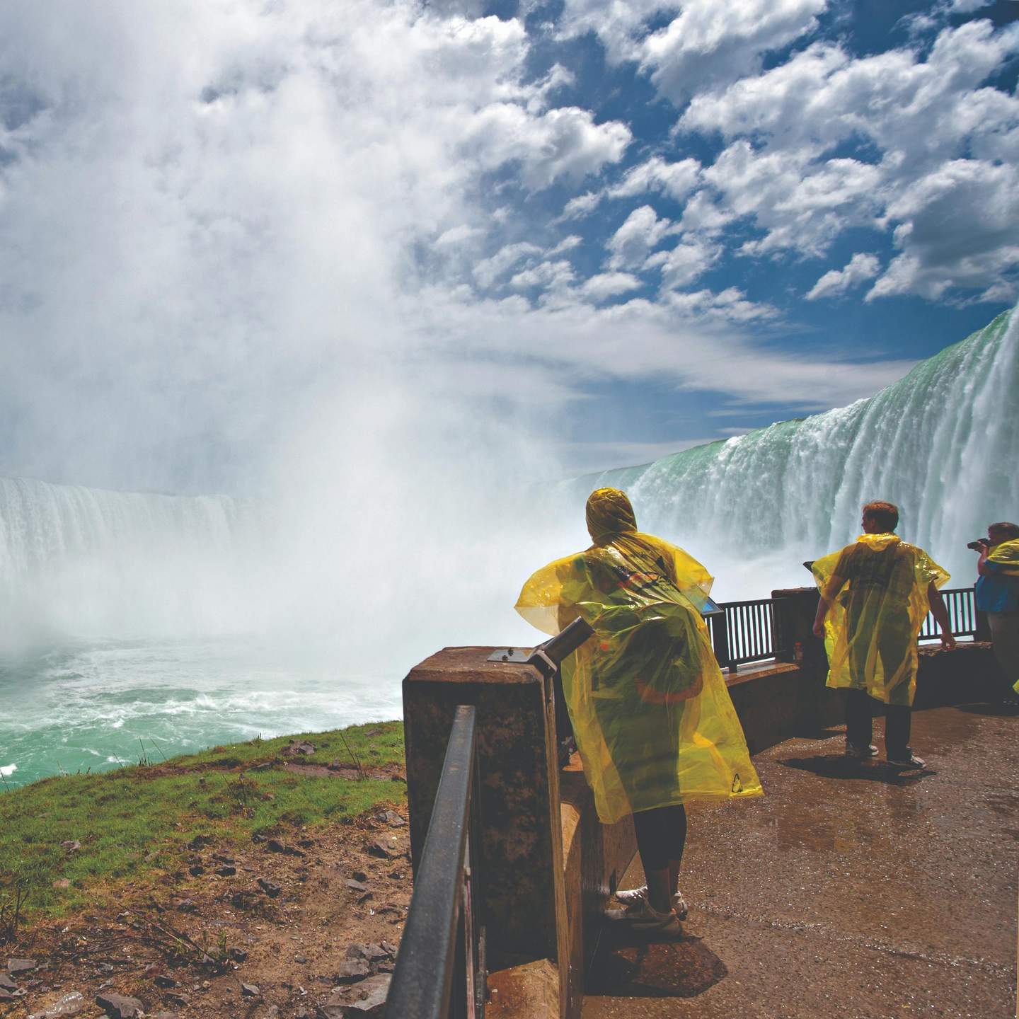 Visitors in yellow rain ponchos standing on a lookout platform near a large, misty waterfall under a partly cloudy sky.