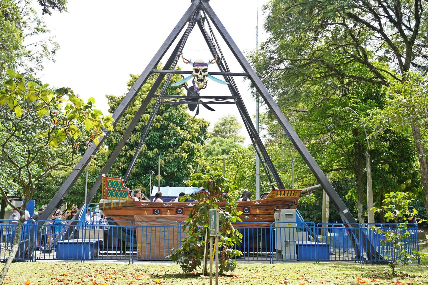Parque Norte Medellín Pirate ship ride at an amusement park with passengers, surrounded by greenery and enclosed by blue metal railings.