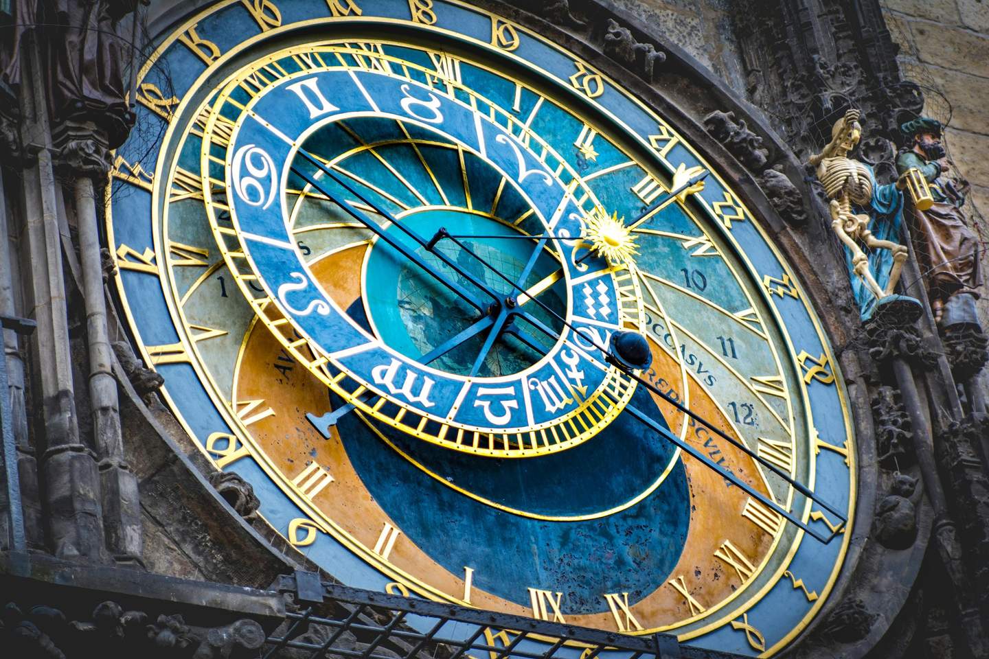 Close-up of an astronomical clock with blue and gold details, Roman numerals, and ornate figures on the sides.