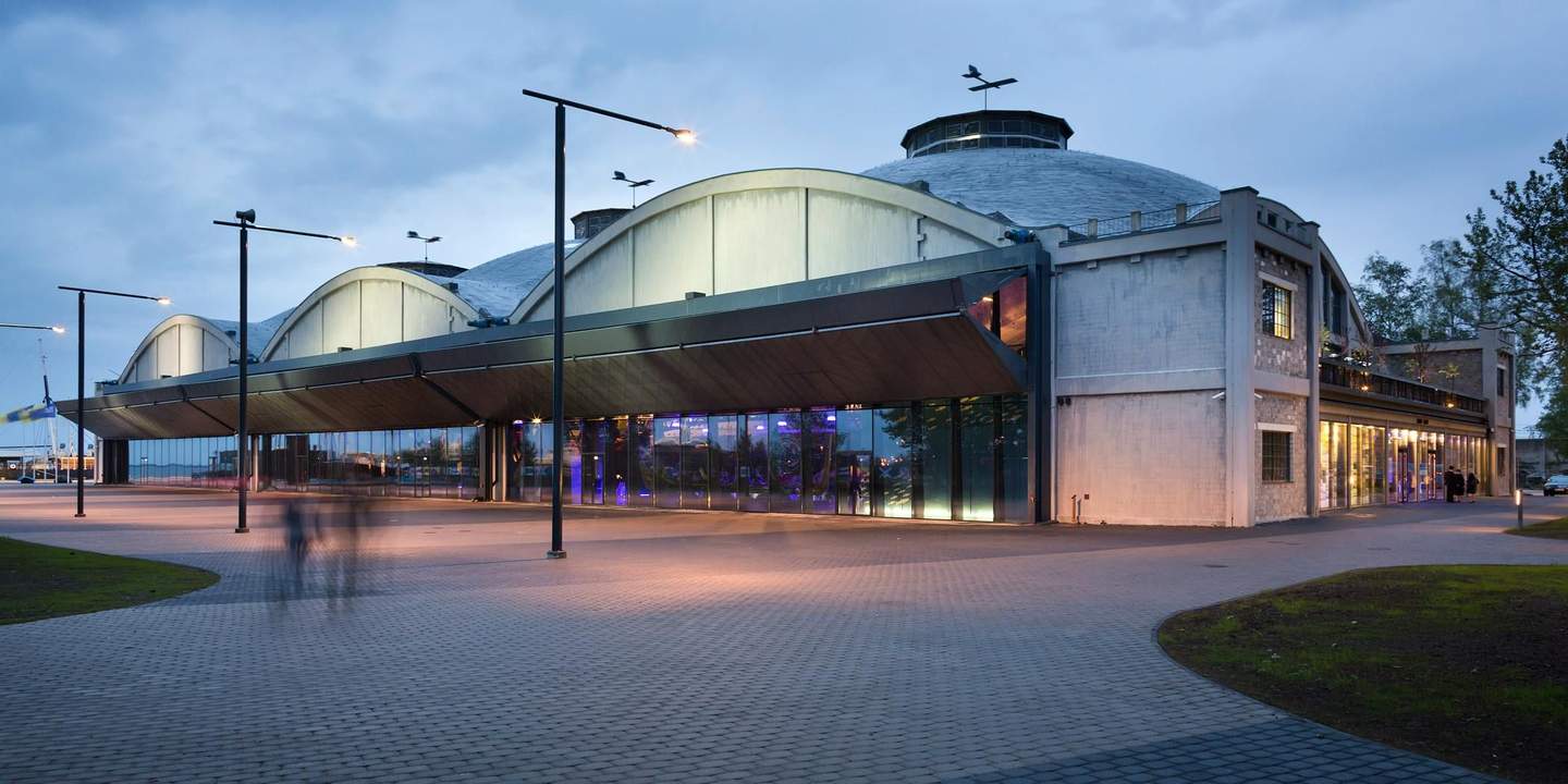 A modern building with large windows and industrial design features, under an evening sky with streetlights and silhouettes of people.