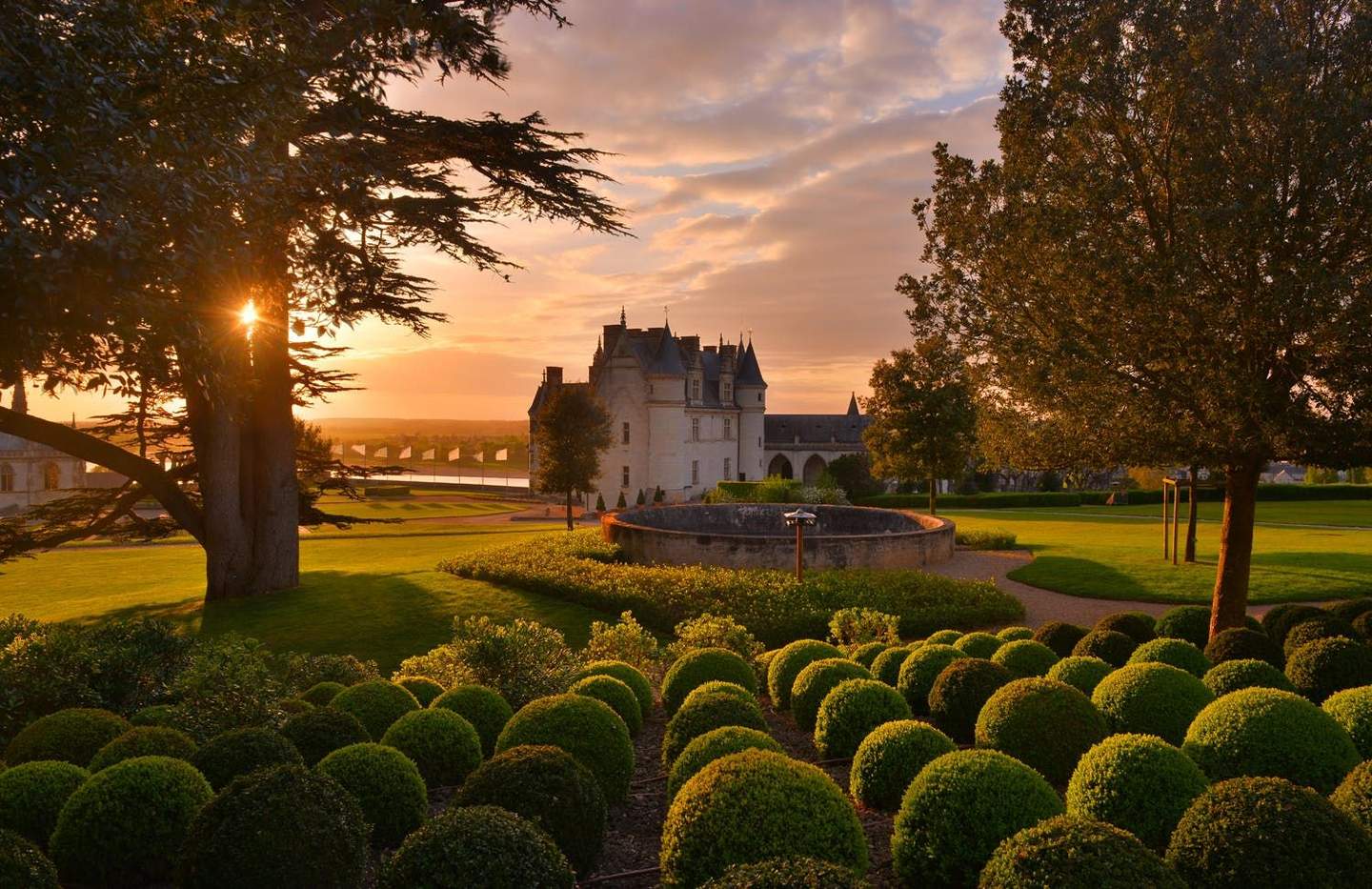 Château d'Amboise A castle at sunset with neatly trimmed bushes, large trees, and a circular stone structure in the garden.