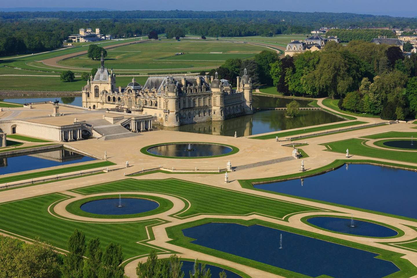 Château de Chantilly Aerial view of an ornate chateau surrounded by manicured lawns and reflecting pools with fountains, situated in a vast green landscape.