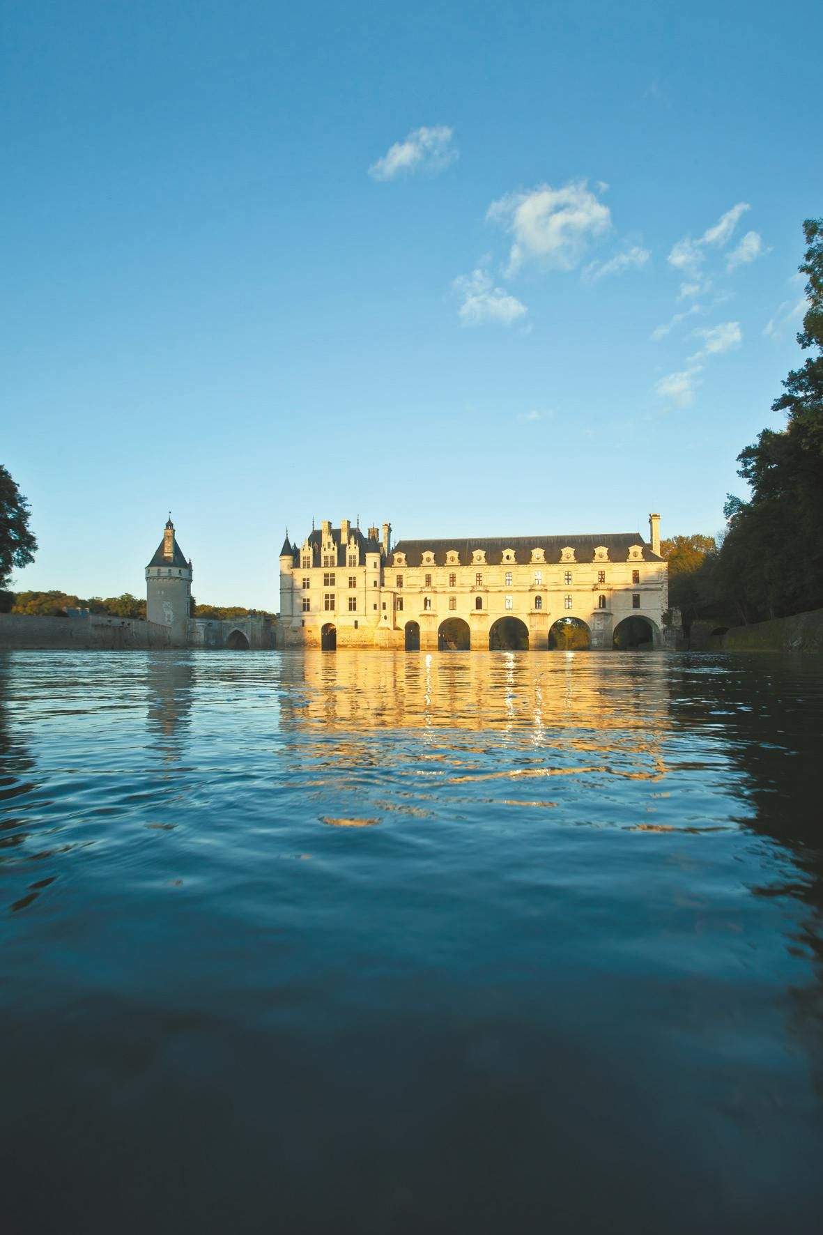 A castle with multiple arches reflected in a calm body of water under a clear blue sky. Trees frame the scene.