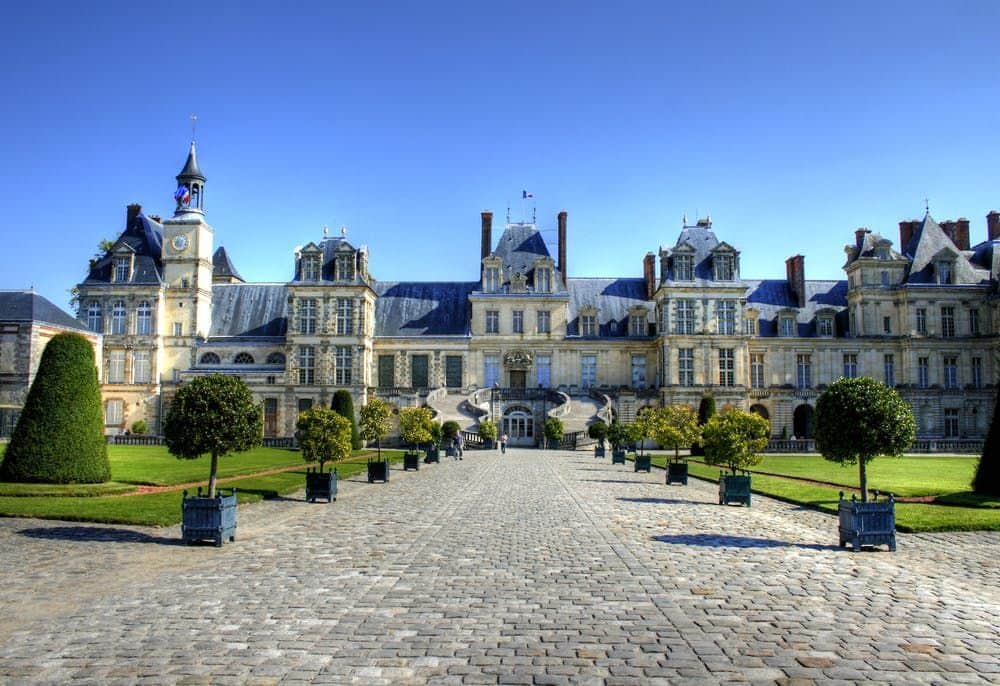 Château de Fontainebleau A large historical building with multiple windows and towers, fronted by a cobblestone path and manicured gardens under a clear blue sky.