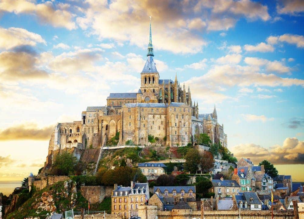 Mont Saint-Michel Abbey A historic stone castle with a tall spire atop a hill overlooks a village under a vibrant, partly cloudy sky.